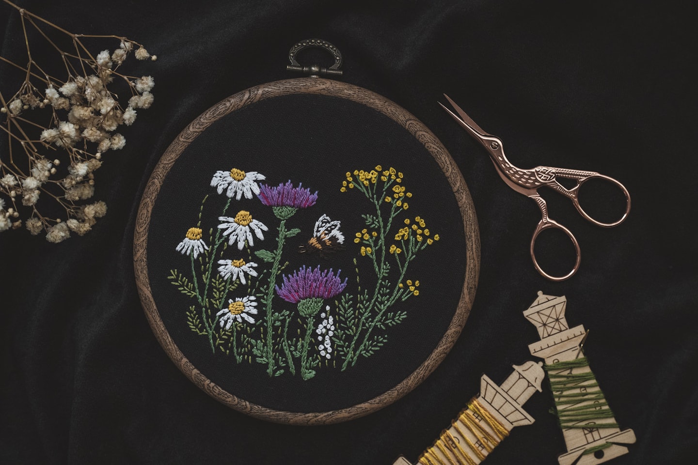 Embroidery hoop with a floral design on black background, between a dried flower and a pair of bird-shaped scissors and two threads.